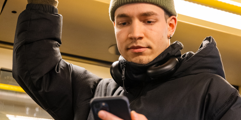 Ein junger Mann mit grüner Mütze und schwarzen Kopfhörern um den Hals steht in einer U-Bahn. Er hält sich mit einer Hand an einer gelben Haltestange fest und blickt konzentriert auf sein Smartphone in der anderen Hand. Auf dem Bild sind Symbole verschiedener Messenger-Apps überlagert, darunter WhatsApp, Telegram, Signal, Facebook Messenger und iMessage, was auf digitale Kommunikation und mobile Vernetzung hindeutet.