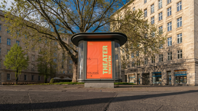 Ein orangefarbenes Veranstaltungsplakat für das Theatertreffen 2026 in einer gläsernen Werbesäule. Das Foto zeigt die Säule auf einem Platz in Berlin, umgeben von historischen Monumentalbauten und Bäumen im Sonnenlicht.