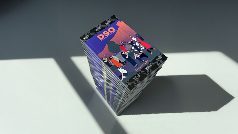 Fotografie eines Stapels von Broschüren des Deutschen Symphonie-Orchesters Berlin (DSO) zur Saison 2025/26. Das Cover zeigt eine farbige Illustration vor der Berliner Philharmonie mit mehreren musizierenden und interagierenden Personen.