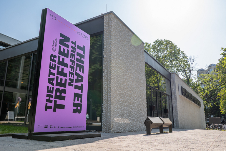 Eine violette Werbestele für das „Theatertreffen“ steht markant im Vordergrund vor der Kieselputz-Fassade des Hauses der Berliner Festspiele. Die Szene ist sonnig, im Hintergrund sind Bäume und die Glasfront des Gebäudes zu sehen.