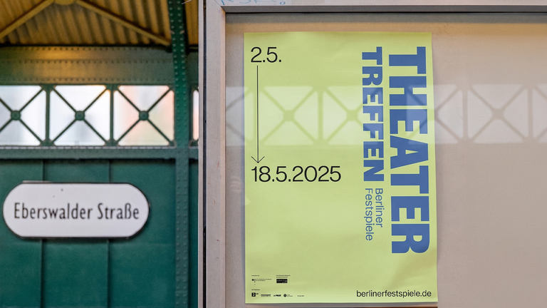 Ein Veranstaltungsplakat für das Theatertreffen 2025 (2.5. bis 18.5.) in Hellgrün und Blau, angebracht in einem Informationskasten. Im unscharfen Hintergrund ist das Stationsschild und die grüne Metallstruktur des U-Bahnhofs Eberswalder Straße in Berlin erkennbar.